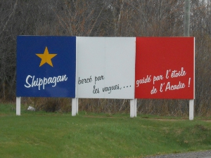 The sign displays, left to right, 'Shippagan' on a blue bar with gold star, 'berce par les vagues...' on the white bar, and 'guide par l'etoile de l'Acadie!' on the red bar.