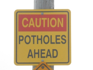 The yellow sign reads 'Caution' with a red background, and then 'Potholes ahead' in black letters