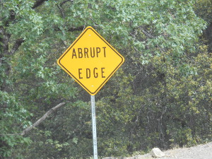 The sign appears immediately next to some trees below the road level at the very edge of the pavement and reads 'ABRUPT EDGE'