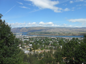 View from the green hills northward, showing the town of The Dalles, the Columbia River as a blue line across the picture, and the plateau beyond, with the sky and clouds above.