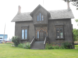 The small but attractive house has two chimneys, a dormered window in the center, and wooden stairs leading up to the front entry.  It is built of dark gray wood.