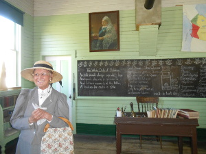 Dressed in a grey suit with a straw hat, the town gossip shares her stories in an old school room