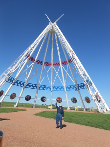 The steel poles which make up the sides of the tepee are painted white, and decorated with lines of red and blue latticework circling around.  The tourist appears to be about one-eighth as tall as the tepee