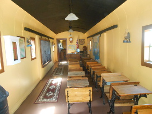 One half of the railroad car has two lines of school desks along one side of the car with an aisle on the other side, and teaching aids mounted on the walls.