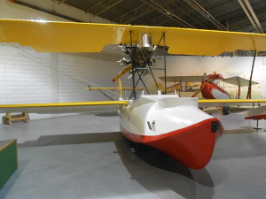 This biplane has bright yellow wings and a pusher propeller high above the red and white boat-shaped cockpit, with a cavity in front for the aerial photographer.