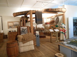 The museum display shows clothes and goods for shipment with a large canoe inverted over the top of the exhibit as if for portaging.