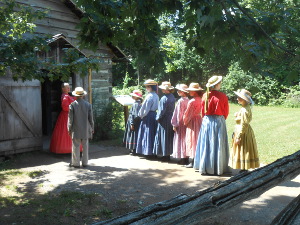 Dressed in brightly colored nineteenth century clothes, schoolgirls and one schoolboy line up for the teacher's inspection of hands prior to entering school.
