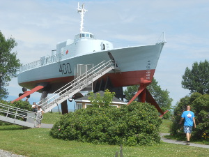 The boat has hydrofoils which are diamond shaped and are resting on the ground, so that the grey Navy boat sits high up in the air, with a staircase leading to it.