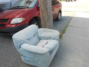 An overstuffed gray easy chair sits on the asphalt in front of the laundry, next to a telephone pole next to a beat up sedan.