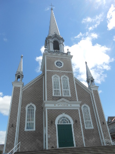 Against the background of a blue sky with white clouds, the church steeple reaches up to the sky, with matching shorter towers on the front corners of the building.