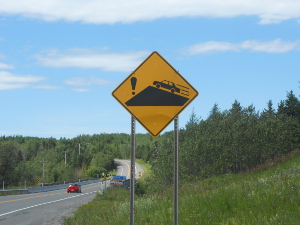 A yellow highway warning sign with an inverted V denoting a hill crest, a car moving rapidly up the right side of the hill, and on the right an exclamation point.
