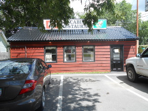 Cars are parked in front of the small dark red restaurant with a sign on the roof.