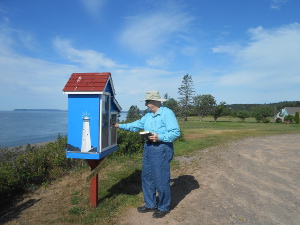Wearing a wide-brimmed Tilley hat, Bob adds books to the Little Free Library mounted on a pole in Port Greville, with a blue sky and the Bay of Fundy in the background.