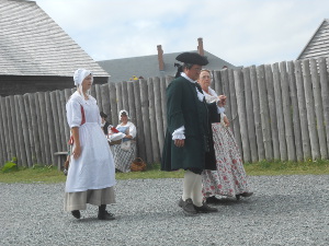 A gentleman in a black surcoat and tri-cornered hat escorts a lady in a floral dress in front of a high wooden fence; a middle-class lady wearing a white apron and bonnet steps behind.