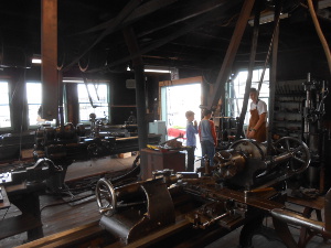 Two boys look up to the tall guide wearing a tan shop apron amidst a shop full of belt-driven lathes and drills.