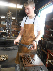 Wearing a white tee shirt under a tan leather apron, the tall, rosy-cheeked high school student is holding a large Crescent wrench