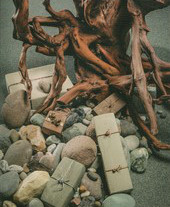 The detail reveals a snarl of bare russet colored driftwood arranged above a trail of rocks, some beach pebbles and some sculpted into rectangular shapes wrapped with metal ribbon and decorations.