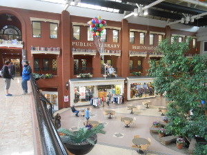 The photograph is taken from a walkway overlooking a large hall on one side of which is the indoor brick facade of the library building, opposite a large tree within the hall.