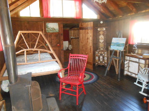 A bed with a handmade wooden frame, an artist's homemade easel, and other creations using local materials to fashion furniture are displayed in a cabin room.