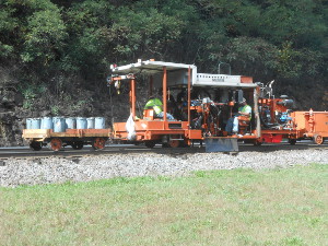 A bright orange maintenance vehicle with three track workers rides the rails, automatically hammering large nails to fasten track to ties