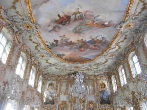 The ballroom is two stories high with upper and lower windows, and a painted ceiling, in white and gold and pastels, brimming with curlicues and baroque ornamentation