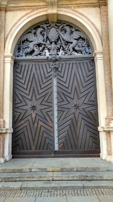 set in a Romanesque arch with the coat of arms in the lintel, the massive dark wooden doors are outlined in octagonal stars