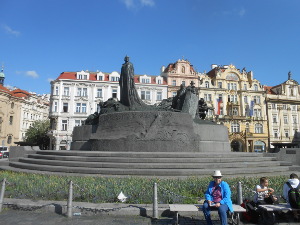 Elsa sits on a bench in front of the huge gray stone monument to Jan Hus