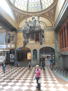 Elsa is standing on the floor of the arcade beneath the suspended statute of Wenceslas sitting astride on the belly of his horse hanging feet up and head down