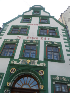 The five story house has white stucco and green and gold windows with German inscriptions and decorative panels and cherubs and the writing painted in dark red, 'Gasthaus Zur Hohen Lilie'