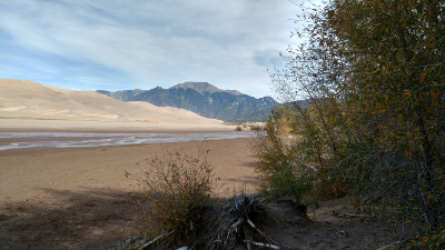 The sand dune appears a light pinkish tan on the left, the mountains in the distance are dark gray, while the foreground is the same sand, darkened in color by the water in the nearly dry creek bed.