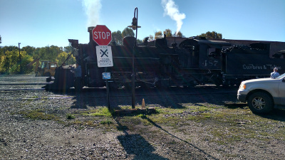A signal light and stop sign in the foreground appear in front of an old black steam locomotive, with a plume of smoke emerging from its stack.