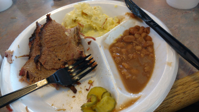 On the styrofoam plate sits a plastic fork, brisket, beans, cole slaw, and pickle