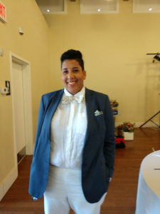 Standing alone in the reception hall, Carmen is facing the camera, wearing a blue jacket and white shirt and pants.