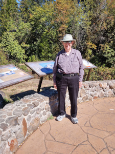Bob stands next to a stone wall wearing jeans and a long sleeved shirt and white walking shoes, with his broad-brimmed Tilly hat protecting him from the sun.