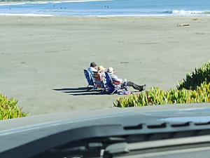 photograph through the window of a vehicle showing three persons sitting in beach chairs on a broad sandy beach with ice plant in the foreground