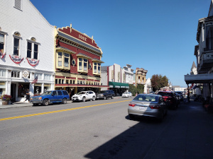 Two story store fronts line this broad street in Ferndale with cars parked on both sides of the street.  The buildings are decorated with Victorian carpentry and decorated with bunting.