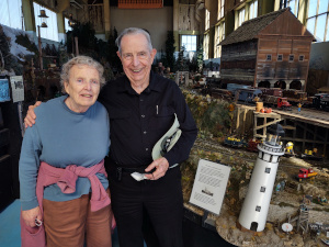 Elsa and Bob had their picture taken inside the building filled with railroad models.  Elsa has her orchid hoodie tied around her waist over her blue long-sleeved shirt while Bob, dressed all in black, has tocked his hat under his arm.