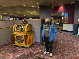 Wearing her blue winter coat and a black mask, Elsa stands in the restaurant lobby next to an old yellow oak Wurlitzer
